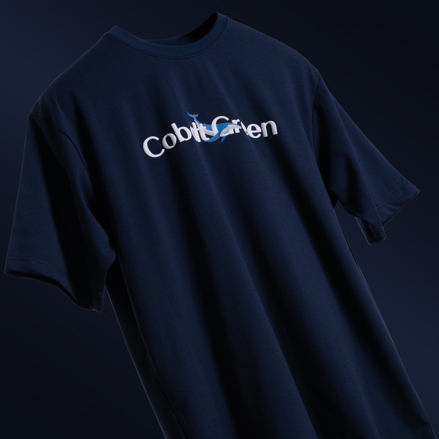 Deep navy blue oversized 'Gonna Be Alright Tee' floating against a matching backdrop, featuring 'Cobalt Green' branding on the front. The back showcases the uplifting phrase 'gonna be alright' with playful blue whale graphics in screen and puff print. Crafted from 220 GSM cotton-rich fabric with bio-washed and gold-finished treatment for a soft, premium feel. The tee's oversized, unisex design offers a relaxed fit, perfect for spreading positivity and comfort.