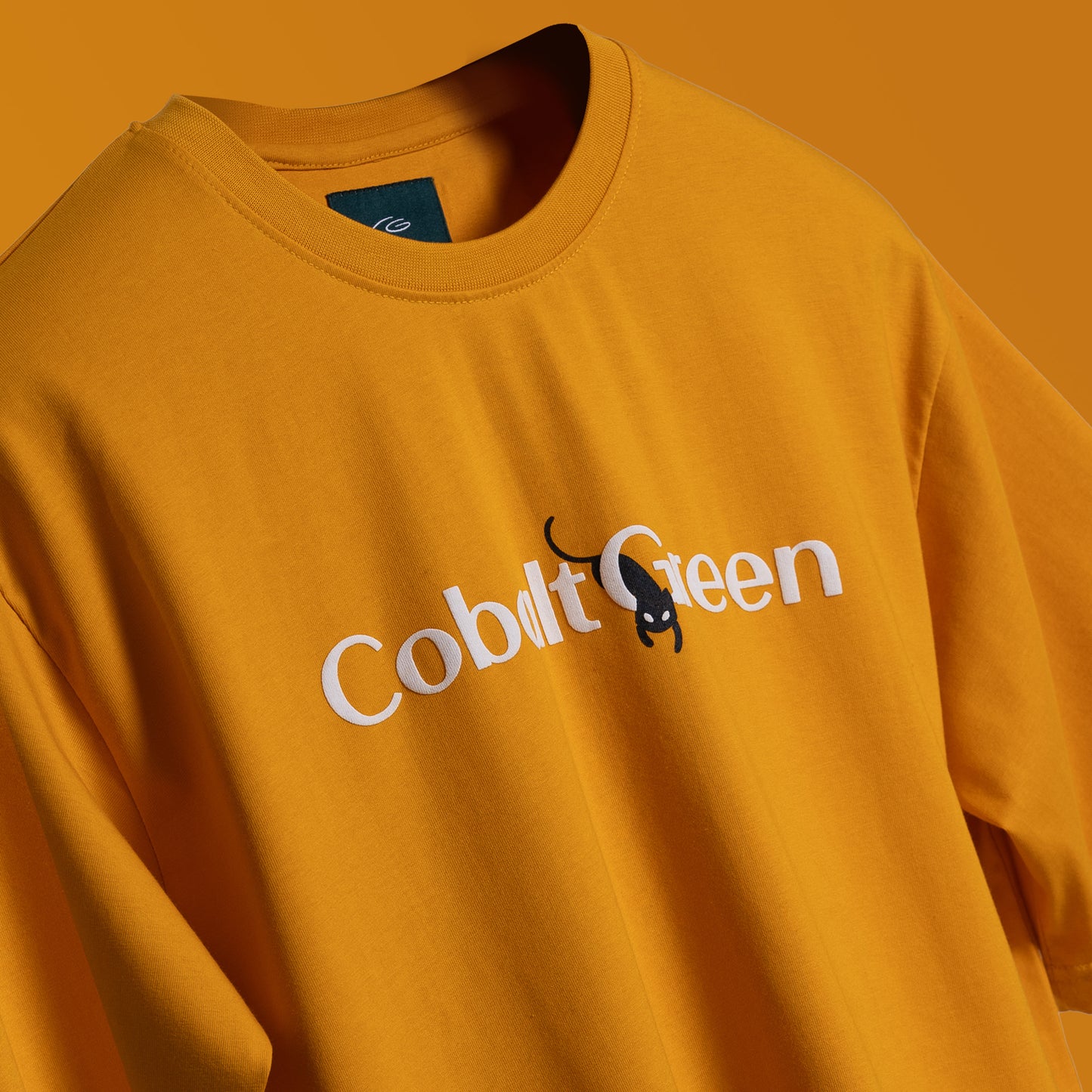 Close up of a Vibrant yellow oversized 'Cat-Not Tee' floating against a clean backdrop, featuring 'Cobalt Green' branding on the front. The back displays playful screen and puff print artwork with the phrase 'I just cat-not stay awake' and a sleepy black cat graphic. Made from 220 GSM cotton-rich fabric, this tee offers a relaxed unisex fit, bio-washed and gold-finished for a soft, premium feel. Perfect for casual wear, lounging, or adding a humorous touch to any outfit.