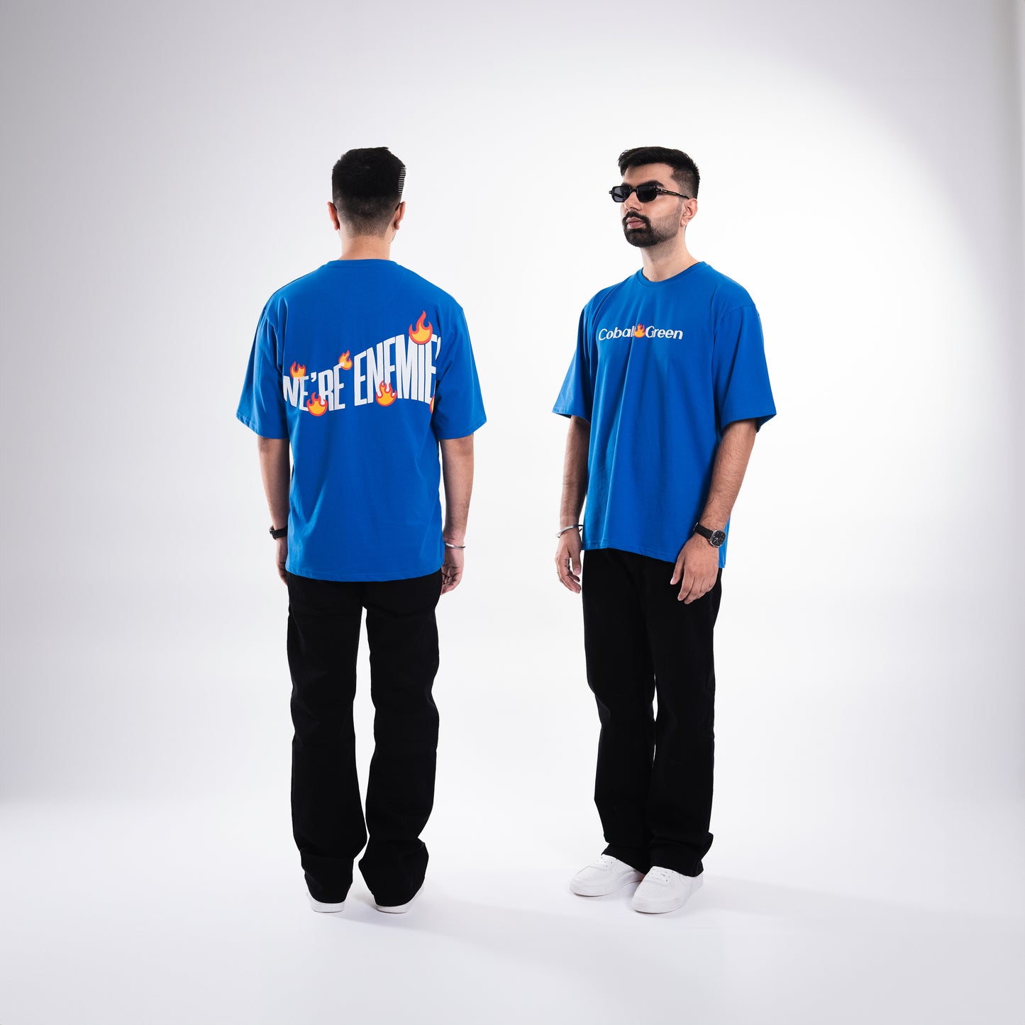 Male 5'10 model wearing the royal blue 'We're Enemies Tee,' posing in a 3/4th view that showcases both the front and back of the tee in the same frame. The front features 'Cobalt Green' branding, while the back displays the bold 'We're Enemies' phrase with dynamic fire symbols in screen and puff print. The oversized, unisex design offers a comfortable and stylish fit, highlighting the tee's bold, edgy aesthetic.