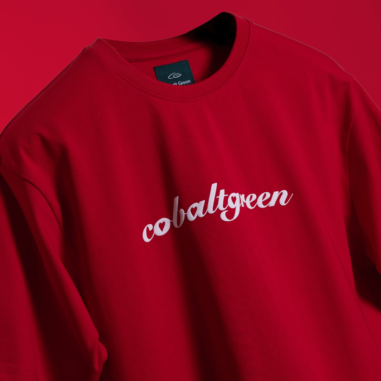 Closeup of Striking red oversized 'Queen's Command Tee' floating against a matching backdrop, featuring 'Cobalt Green' branding on the front. The back showcases the bold 'Queen's Command' phrase with an intricately illustrated Queen of Hearts card in screen and puff print. Crafted from 220 GSM cotton-rich fabric with bio-washed and gold-finished treatment for a soft, premium feel. The oversized, unisex design offers a relaxed, flattering fit, perfect for expressing confidence and royal style.