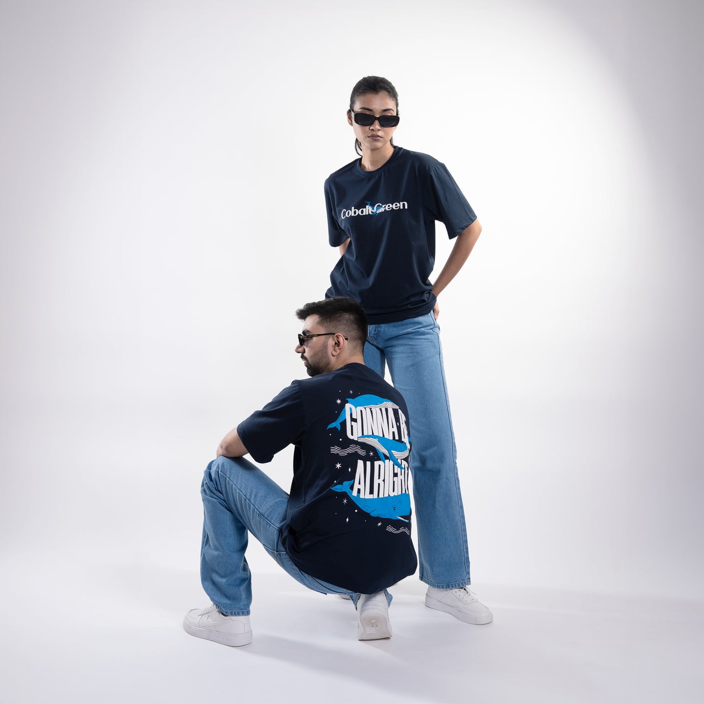 Male and female models, both 5'10", wearing the deep navy blue 'Gonna Be Alright Tee' in a posed shot. The front features 'Cobalt Green' branding, while the back showcases the uplifting 'gonna be alright' phrase with playful blue whale graphics. The oversized fit and unisex design create a relaxed, stylish look that flatters both body types, emphasizing comfort and positive vibes.