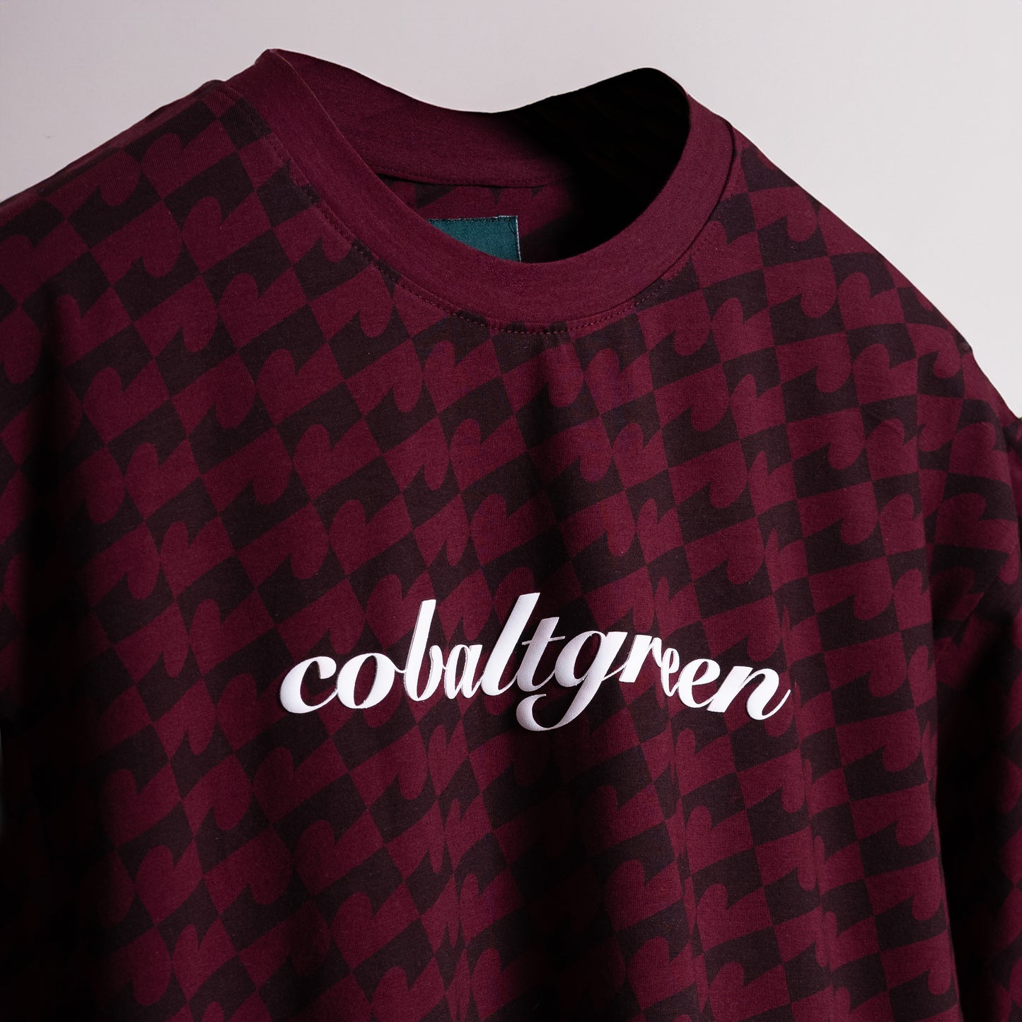 Close-up of a Maroon oversized t-shirt with an all-over-print design, floating against a neutral background for a tone-on-tone effect. The shirt features 'Cobalt green' branding on the chest plate, creating a subtle yet stylish look that emphasizes the seamless blend of color and design.