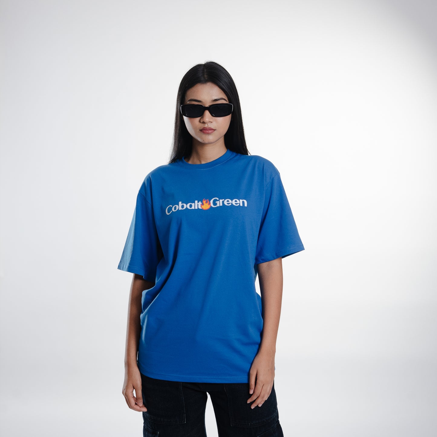 Female model wearing the royal blue 'We're Enemies Tee' in a mid shot, showcasing the oversized fit. The front features the 'Cobalt Green' branding, while the back displays the bold 'We're Enemies' phrase with dynamic fire symbols. The relaxed, unisex design provides a comfortable and flattering look, perfect for making a bold style statement.