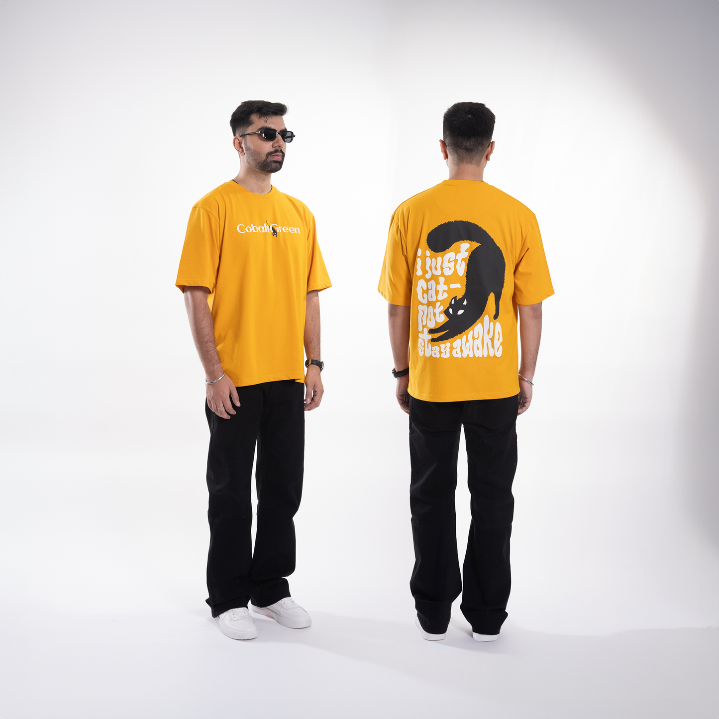 Male model, 5'10", wearing the vibrant yellow 'Cat-Not Tee' in a split frame showing both 3/4th and back angles. The front displays 'Cobalt Green' branding, while the back features a playful 'I just cat-not stay awake' phrase with a sleepy black cat graphic. The oversized fit and unisex design offer a relaxed, comfortable look, perfect for casual styling.