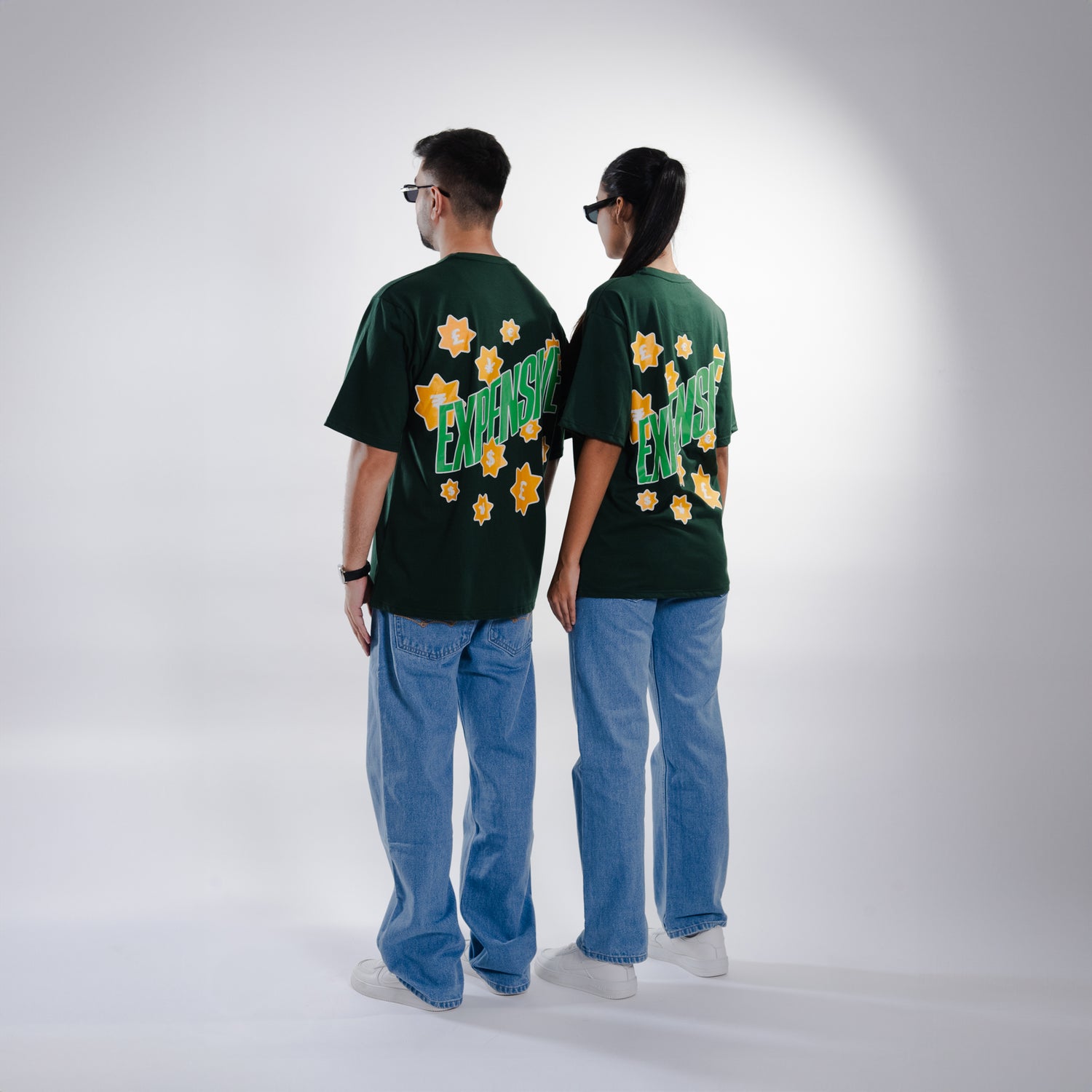 Male and female models, both 5'10", wearing the deep dark green 'Expensive Tee' in a back shot. The oversized fit is showcased as the back features bold 'Expensive' screen and puff print, accented with currency symbols for a luxe touch. The tee’s relaxed, unisex design flatters both body types, offering a stylish and comfortable look.