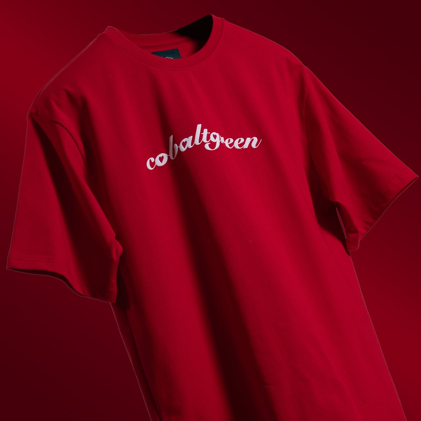 Striking red oversized 'Queen's Command Tee' floating against a matching backdrop, featuring 'Cobalt Green' branding on the front. The back showcases the bold 'Queen's Command' phrase with an intricately illustrated Queen of Hearts card in screen and puff print. Crafted from 220 GSM cotton-rich fabric with bio-washed and gold-finished treatment for a soft, premium feel. The oversized, unisex design offers a relaxed, flattering fit, perfect for expressing confidence and royal style.