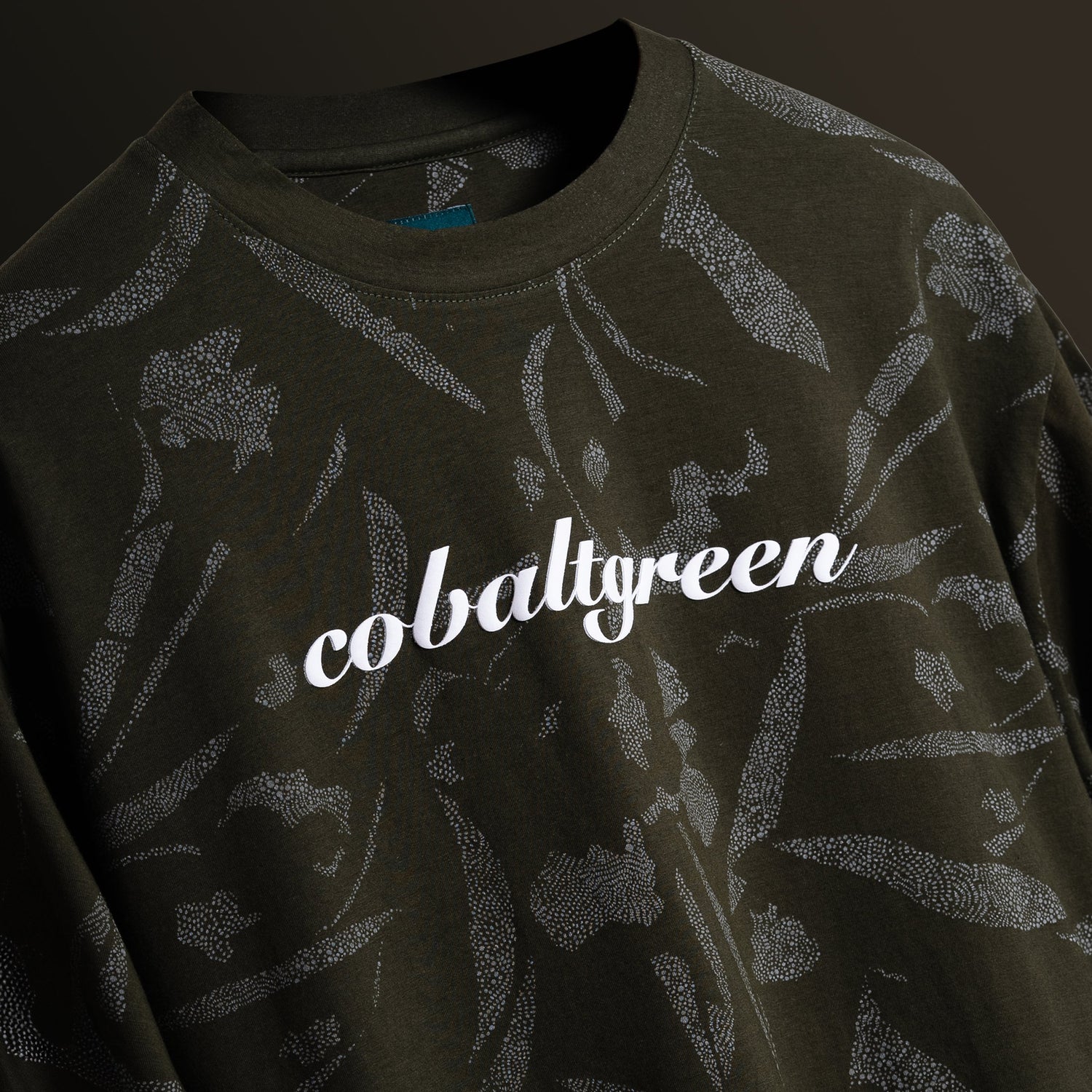 Olive oversized t-shirt with an all-over-print design, floating against a matching olive-toned background for a cohesive, monochromatic effect. The shirt is accented with 'Cobalt green' branding on the chest plate, offering a sleek and modern look.