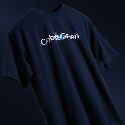 Deep navy blue oversized 'Gonna Be Alright Tee' floating against a matching backdrop, featuring 'Cobalt Green' branding on the front. The back showcases the uplifting phrase 'gonna be alright' with playful blue whale graphics in screen and puff print. Crafted from 220 GSM cotton-rich fabric with bio-washed and gold-finished treatment for a soft, premium feel. The tee's oversized, unisex design offers a relaxed fit, perfect for spreading positivity and comfort.
