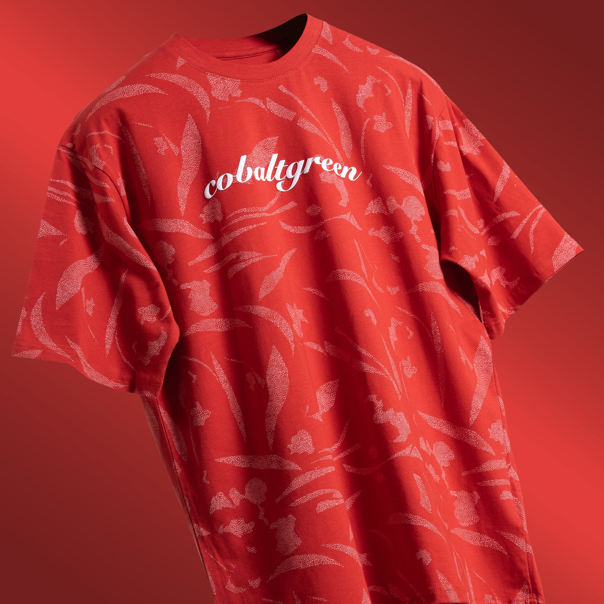 Brick-red oversized t-shirt with an all-over-print design, floating against a clean background. The shirt features 'Cobalt green' branding prominently on the chest plate, highlighting its unique style and bold color. Perfect for a relaxed yet fashion-forward look.