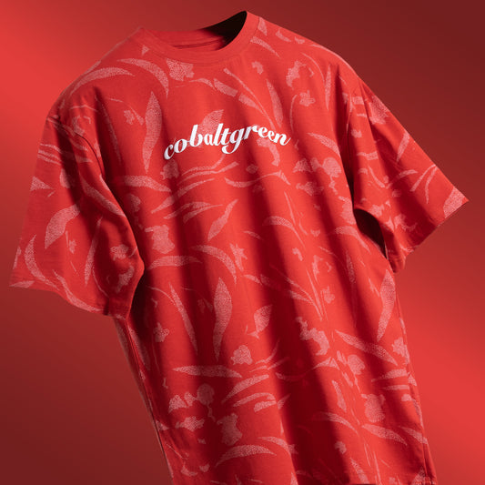 Brick-red oversized t-shirt with an all-over-print design, floating against a clean background. The shirt features 'Cobalt green' branding prominently on the chest plate, highlighting its unique style and bold color. Perfect for a relaxed yet fashion-forward look.