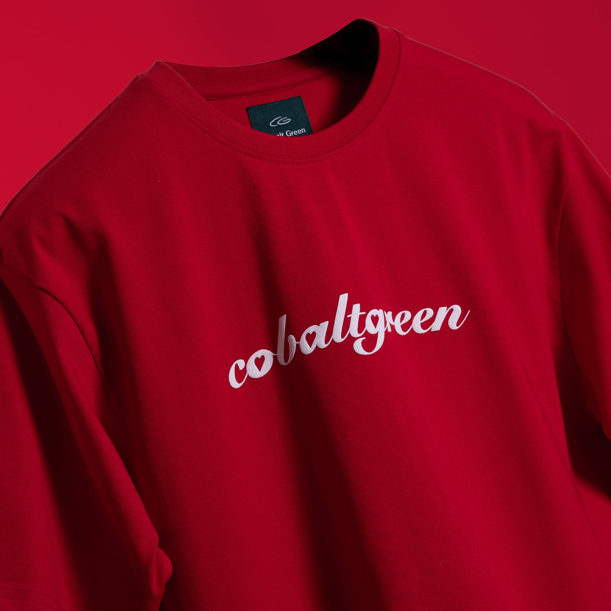 Closeup of Striking red oversized 'Queen's Command Tee' floating against a matching backdrop, featuring 'Cobalt Green' branding on the front. The back showcases the bold 'Queen's Command' phrase with an intricately illustrated Queen of Hearts card in screen and puff print. Crafted from 220 GSM cotton-rich fabric with bio-washed and gold-finished treatment for a soft, premium feel. The oversized, unisex design offers a relaxed, flattering fit, perfect for expressing confidence and royal style.