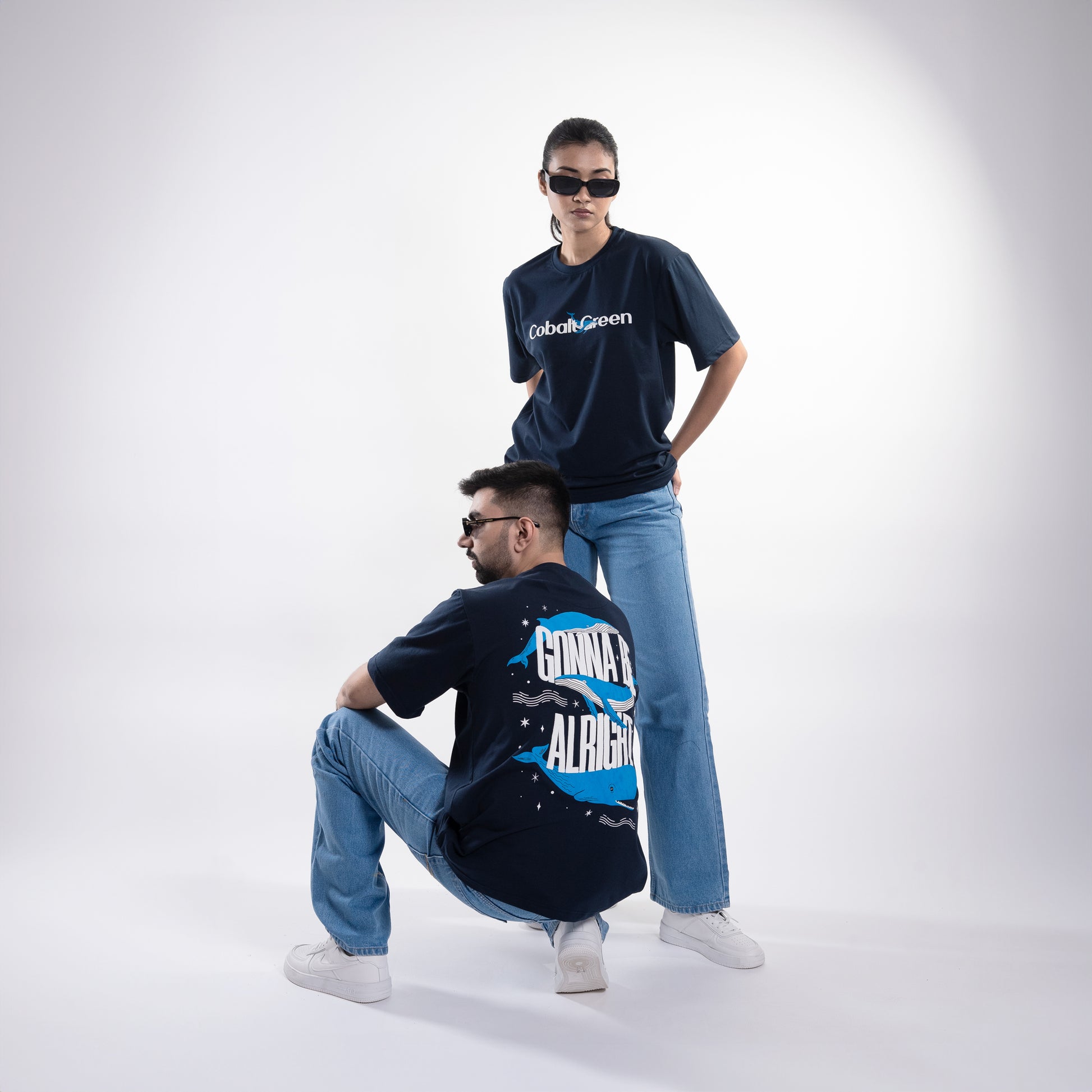 Male and female models, both 5'10", wearing the deep navy blue 'Gonna Be Alright Tee' in a posed shot. The front features 'Cobalt Green' branding, while the back showcases the uplifting 'gonna be alright' phrase with playful blue whale graphics. The oversized fit and unisex design create a relaxed, stylish look that flatters both body types, emphasizing comfort and positive vibes.