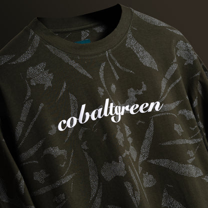 Close up of an Olive oversized t-shirt with an all-over-print design, floating against a matching olive-toned background for a cohesive, monochromatic effect. The shirt is accented with 'Cobalt green' branding on the chest plate, offering a sleek and modern look.