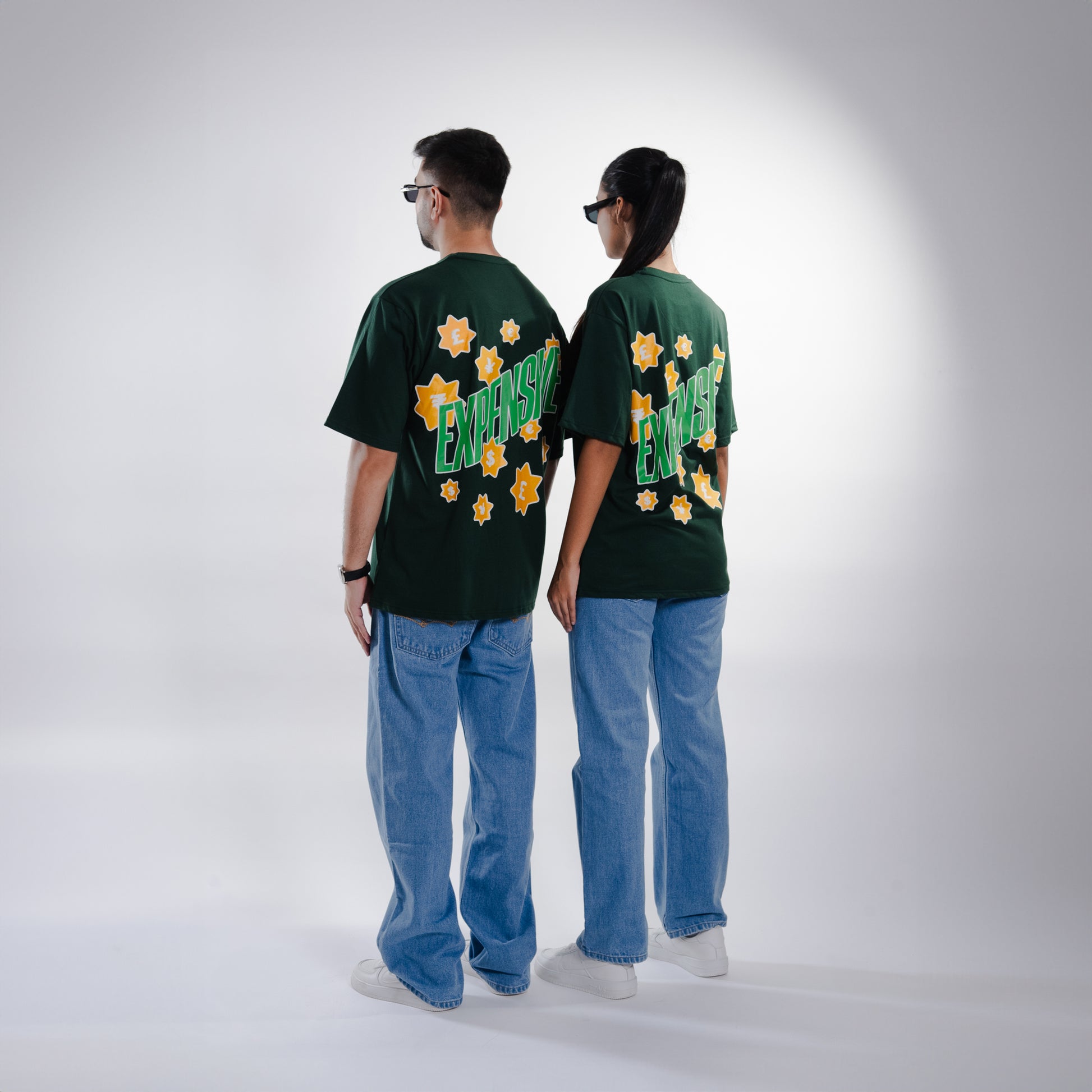 Male and female models, both 5'10", wearing the deep dark green 'Expensive Tee' in a back shot. The oversized fit is showcased as the back features bold 'Expensive' screen and puff print, accented with currency symbols for a luxe touch. The tee’s relaxed, unisex design flatters both body types, offering a stylish and comfortable look.