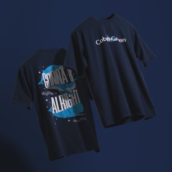Video showcasing two oversized 'Gonna Be Alright Tees' floating against a matching deep navy blue backdrop, rotating to display both the front and back. The front features 'Cobalt Green' branding, while the back highlights the uplifting 'gonna be alright' phrase and playful blue whale graphics in screen and puff print. The video emphasizes the premium, oversized fit and positive vibe of these unisex tees.